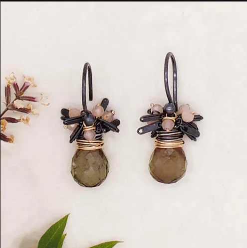 Pair of earrings on white background. Hooks of earrings are black with black and peach moonstone fringe pieces coming from top. Gold colored wire wraps around underneath fringe and connects to amber colored round gem stone