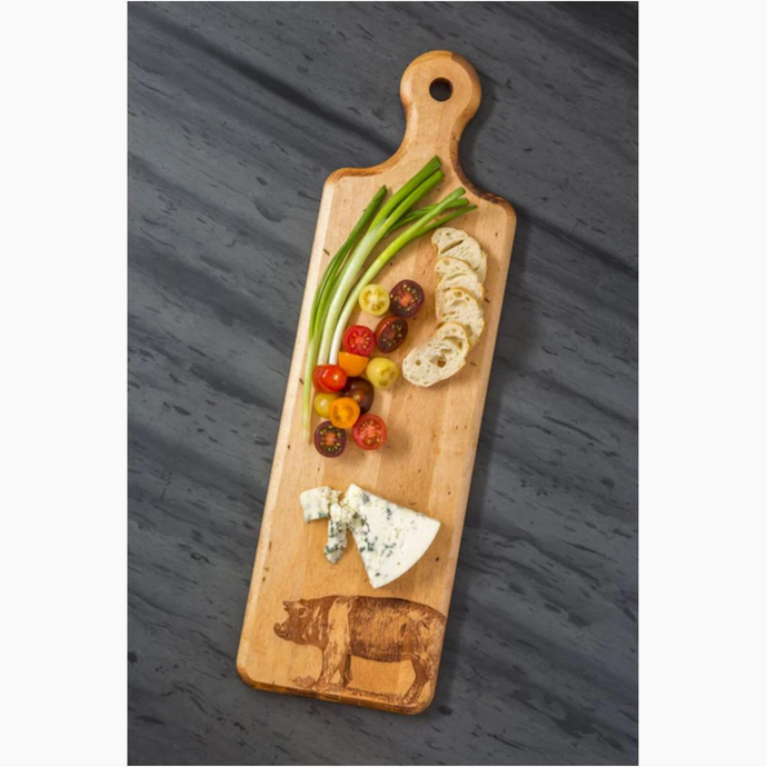 Image of long wooden cutting board on dark gray background. Image of a pig burned into bottom right corner of board with rounded handle at other end. Rest of board covered in cheese, bread slices, tomatoes, and green onions.