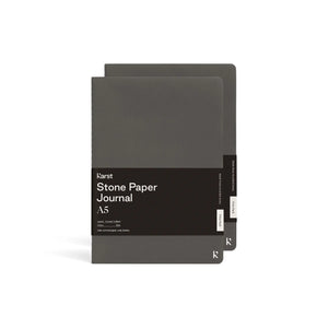 Two dark gray journals stagger stacked on top of each other; Front journal has large black rectangular paper wrap half way down reads "KARST" "Stone paper Journal" "A5" on right side; "K" printed in black on bottom left corner of journal cover