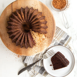 Baked dark brown bundt cake with light brown powder on top sitting on wooden platter; Slice of cake on white plate next to platter with fork next to cake and brown/gray checkered napkin under plate; Small sifter with light brown powder topping in top left corner